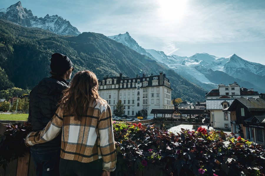 A couple look out over a town at the foot of a mountain range in Europe. 